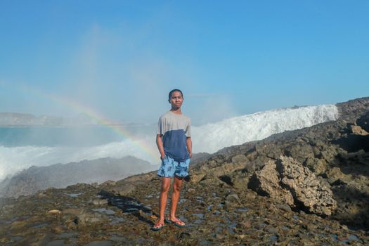 A man stands on a rocky shore and the waves crash against a cliff. Rainbow phenomenon in water fog. Waves hitting round rocks and splashing.