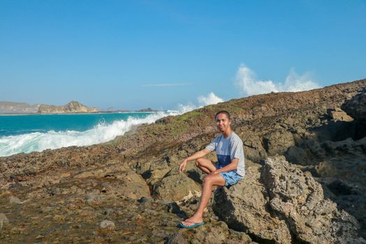 young man in a T-shirt and blue shorts sitting on a rocky shore. This is a view of man sitting on the rock by the sea.