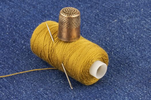 On a blue denim lies reel yellow thread with a needle and a metal copper thimble