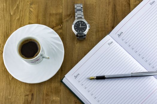 On a wooden surface lies an open diary with a pen, a watch with a bracelet and a cup of coffee costs