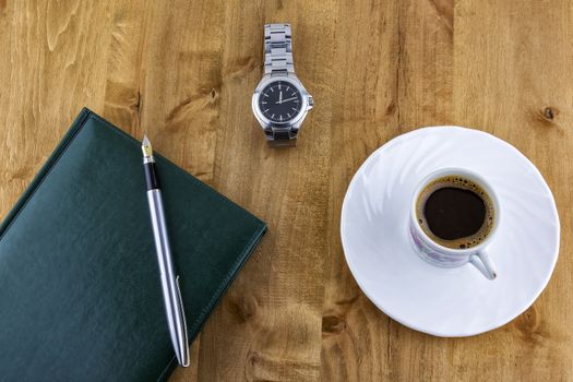 On a wooden surface lies diary with a pen, a watch with a bracelet and a cup of coffee costs