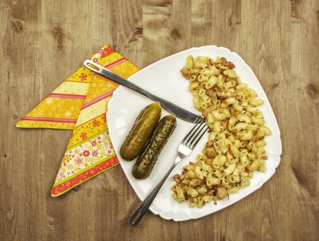 On a wooden surface is white dish with pasta, fried meat and onions and pickles