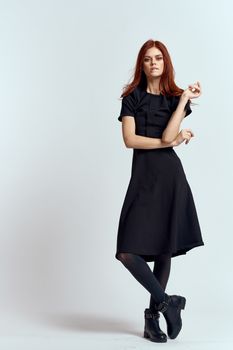 A woman in a black dress on a light background and pantyhose shoes red hair and pose in full growth. High quality photo