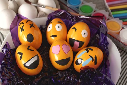Emoticons Easter Eggs on purple cellophane paper. More white eggs, brush and paints in the background.