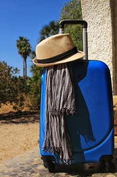 Suitcase prepared for the holidays with striped foulard and white hat. Palm tree and blue sky in the background