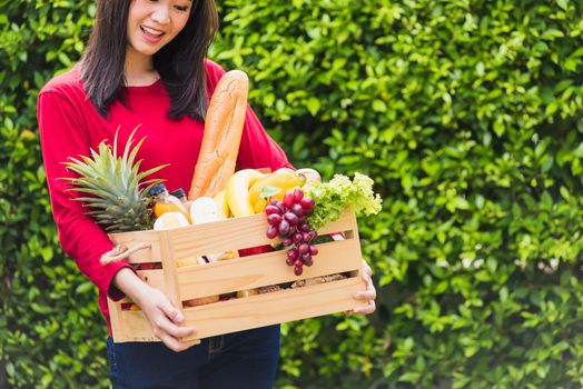 Portrait of Asian beautiful young woman farmer standing she smile and holding full fresh food raw vegetables fruit in a wood box in her hands on green leaves background