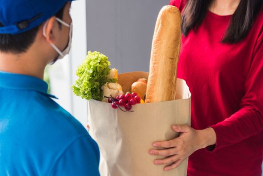 Asian young delivery man in uniform wear protective face mask he making grocery service giving fresh food to woman customer receiving front house under pandemic coronavirus, Back to new normal concept