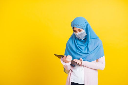 Asian Muslim Arab, Portrait of happy beautiful young woman Islam religious wear veil hijab and face mask protect she quarantines disease coronavirus touch on tablet screen isolated yellow background