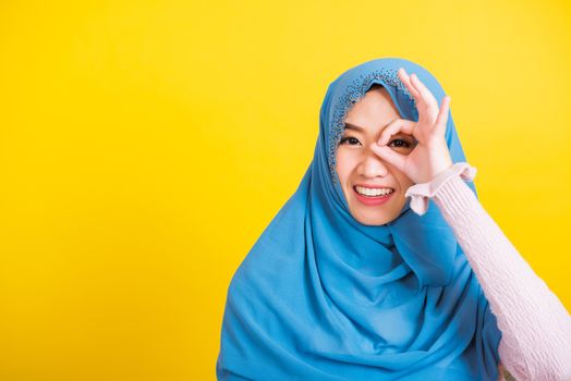 Asian Muslim Arab, Portrait of happy beautiful young woman Islam religious wear veil hijab funny smile she show gesture fingers in okay gesture symbol, OK sign over her eyes isolated yellow background