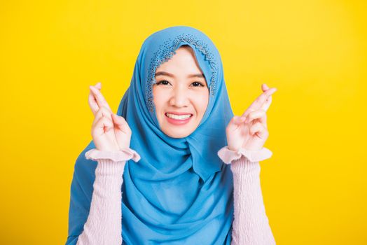 Asian Muslim Arab, Portrait of happy Asian beautiful young woman Islamic religious wear veil hijab she smiling and holding fingers crossed for good luck, studio shot isolated on yellow background