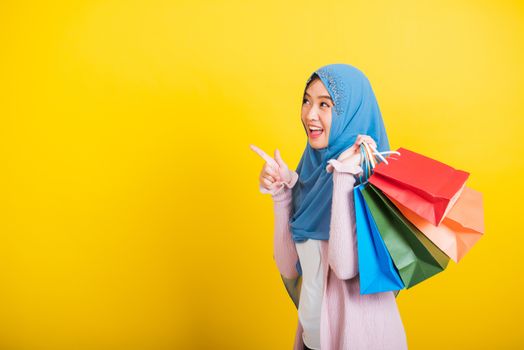 Asian Muslim Arab, Portrait of happy beautiful young woman Islam religious wear veil hijab funny smile she holding colorful shopping bags, studio shot isolated on yellow background