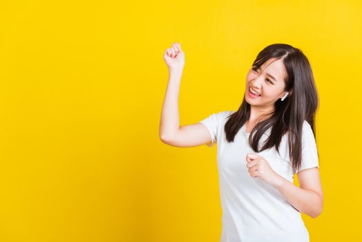 Portrait Asian of a happy beautiful young woman wear wireless earphone listening to music from smartphone having fun and dancing to sounds of favorite music studio shot isolated on yellow background