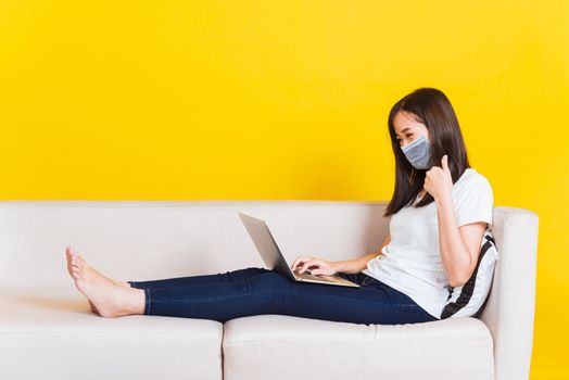 Portrait Asian of beautiful young woman sitting on sofa wearing medical face mask protective she work from home with laptop computer during Coronavirus studio shot isolated on yellow background