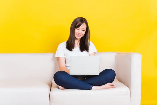 Portrait Asian of happy beautiful young woman work from home she sitting on sofa using laptop computer in house living room studio shot isolated on yellow background