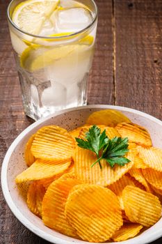 Fresh ice cold water drink with lemon near to fried corrugated golden potato chips with parsley leaf in wooden bowl on wood backdrop, angle view