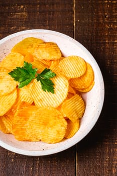 Fried corrugated golden potato chips with parlsey leaf in wooden bowl on wood backdrop, top view