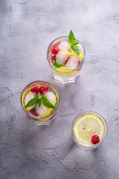 Fresh cold ice water drink with lemon, raspberry fruits and mint leaf in three glass on stone concrete background, summer diet beverage, angle view selective focus