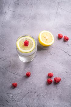 Fresh cold sparkling water drink with lemon, raspberry fruits in glass on stone concrete background, summer diet beverage, angle view selective focus