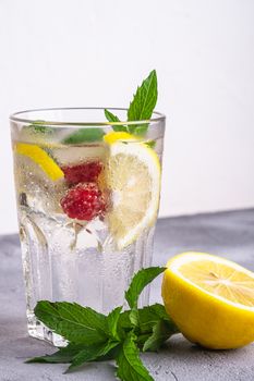 Fresh cold ice water drink with lemon, raspberry fruits and mint leaf in faceted glass on stone concrete background, summer diet beverage, angle view