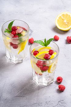 Fresh cold ice water drink with lemon, raspberry fruits and mint leaf in two faceted glass on stone concrete background, summer diet beverage, angle view