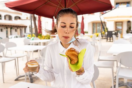 Beautiful fashionable blonde woman has breakfast outdoors in the empty dehors of a bar with a medical mask lowered on her face to eat brioche and drink espresso coffee with milk or cappuccino