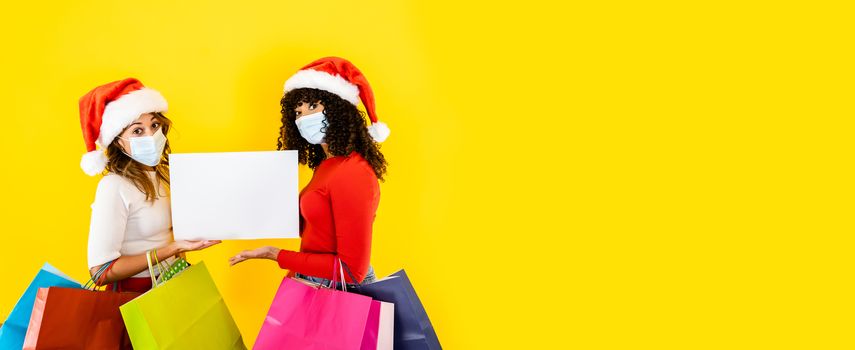 Two young cute women wearing Santa Claus hat looking to the camera with wide eyes and medical mask, holding sign for copy space with many shopping bags in arms - Christmas banner for Coronavirus