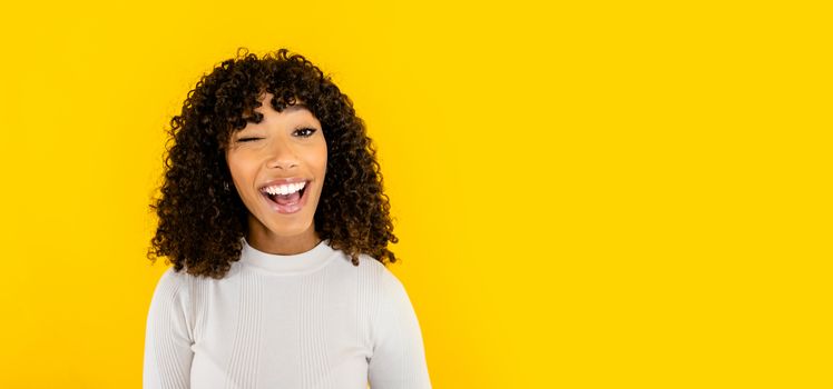 Black Hispanic curly beautiful young woman casual dressed winks looking at the camera smiling with open mouth on big yellow background copy space - One female optimistic person laughing