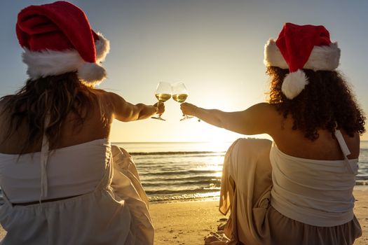 Rear view of two multiethnic best female friends wearing Santa hat sitting on the seashore and toasting with a glass of white wine in the sunset or dawn - Scene of female people celebrating holidays