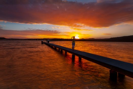 Silhouette figure standing on the jetty taking photos of amazing vivid red sunsets,
