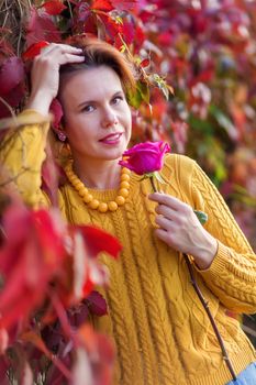Portrait of 30 year old woman holding rose and standing near bush of red wild vine leaves in autumn park looking at the camera