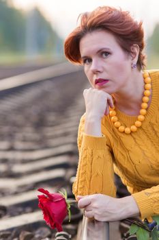 Portrait of beautiful 30 year old woman sitting on rails of the railway holding red rose