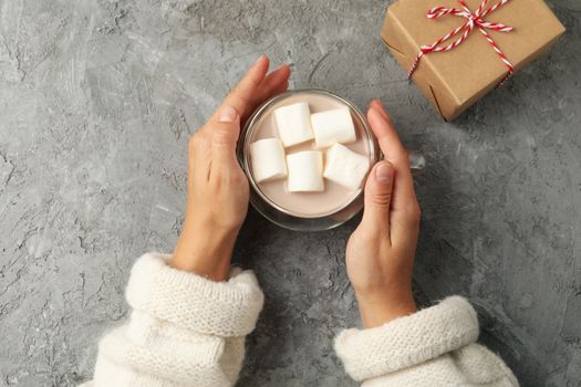 Female hands hold cup of coffee with marshmallows on gray background, top view