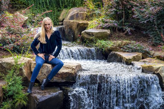 A gorgeous blonde model sits near a waterfall at a local park