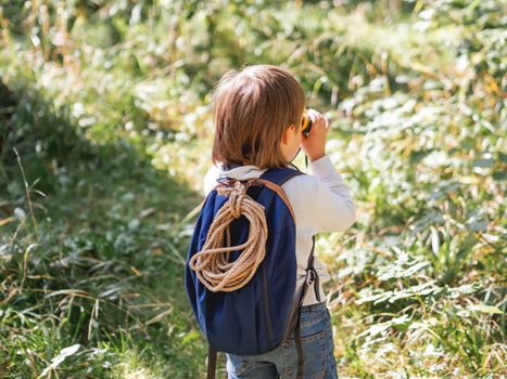 Curious boy is hiking in forest. Outdoor leisure activity for kids. Child looks through binoculars on tree foliage. Sunny day at autumn or summer day.