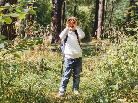 Curious boy is hiking in forest. Outdoor leisure activity for kids. Child looks through binoculars on tree foliage. Sunny day at autumn or summer day.