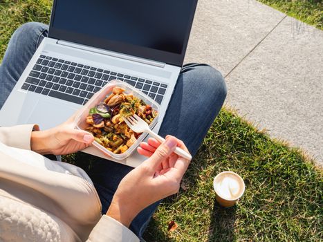 Woman sits on park bench with laptop and take away lunch box, cardboard cup of coffee. Healthy bowl with vegetables. Casual clothes, urban lifestyle of millennials. Healthy nutrition.