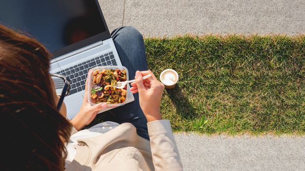 Woman sits on park bench with laptop and take away lunch box, cardboard cup of coffee. Healthy bowl with vegetables. Casual clothes, urban lifestyle of millennials. Healthy nutrition. Copy space.