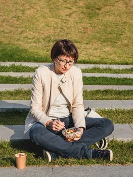 Woman sits on park bench with take away lunch box, cardboard cup of coffee. Healthy bowl with vegetables. Casual clothes, urban lifestyle of millennials. Healthy nutrition.