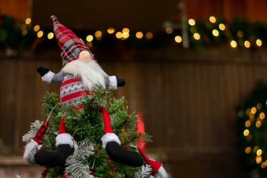 A Christmas Gnome Dressed in a Red Sweater and Hat With a Big Nose on Top of a Christmas Tree With Bokeh Christmas Lights in the Background