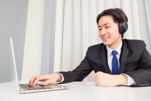 Work from home, Asian young businessman smile wearing headphones and suit video conference call or facetime with laptop computer on desk at home office