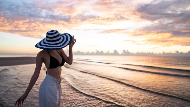 Woman in top bikini and white long pant wearing hat on the beach with a beautiful sunrise or sunset in background.