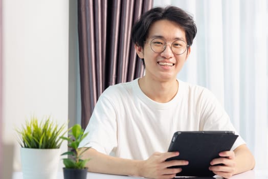 Happy Asian young business handsome man work from home office wear glasses, t-shirt comfortable he smiling and using a black modern smart digital tablet computer to read email or learning on desk