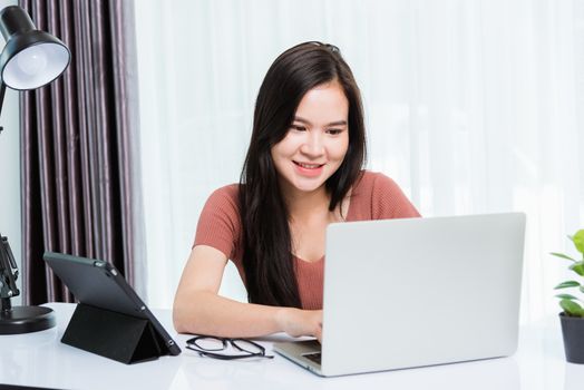 Work from home, Smiling Happy Asian business young beautiful woman sitting on desk workspace use modern smart tablet and laptop computer technology video call conferencing to team at home office