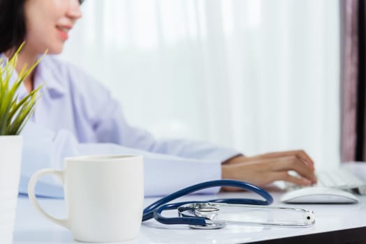 Asian beautiful female doctor smiling wearing doctor's uniform with stethoscope typing on keyboard desktop computer looking to monitor on the desk at hospital office, Medical healthcare concept