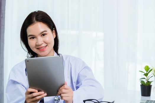 Asian young beautiful woman doctor on desk front laptop computer she holding modern smart digital tablet on hand during quarantines disease coronavirus or COVID-19 at hospital office
