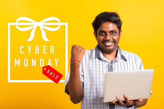 Asian happy young black man excited holding laptop computer clenching fists and raising a hand for winner sign his success with Cyber Monday text in gift box on side, isolated on yellow background