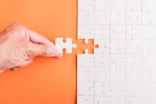 Top view flat lay of hand-holding last piece white paper jigsaw puzzle game last pieces put to place for solve problem complete mission, studio shot on an orange background, quiz calculation concept