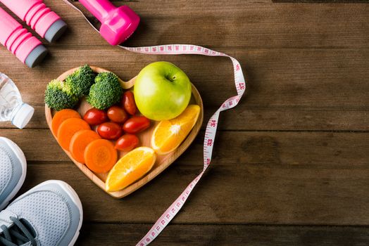 Top view of fresh fruits and vegetables in heart plate wood (apple, carrot, tomato, orange, broccoli) and dumbbells, sport shoes sports equipment on wooden table, Healthy lifestyle diet food concept