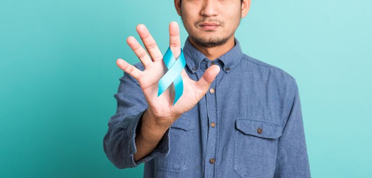 Asian portrait happy handsome man posing he holding light blue ribbon for supporting people living and illness, studio shot isolated on blue background, Prostate Cancer Awareness in November concept