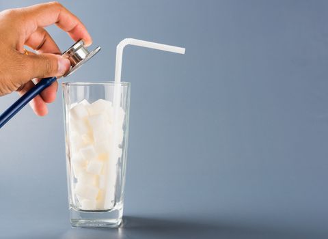 Hand of doctor hold stethoscope check on glass full of white sugar cube sweet food ingredient, isolated on gray background, health high blood risk of diabetes and calorie intake unhealthy drink